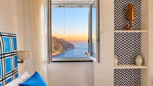A bedroom with a bed, a window with a view of a coastline, a wall with a chevron pattern, and a shelf with decorative items. - Gala Residence Villa Giovanna (Ravello)
