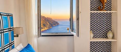 A bedroom with a bed, a window with a view of a coastline, a wall with a chevron pattern, and a shelf with decorative items.