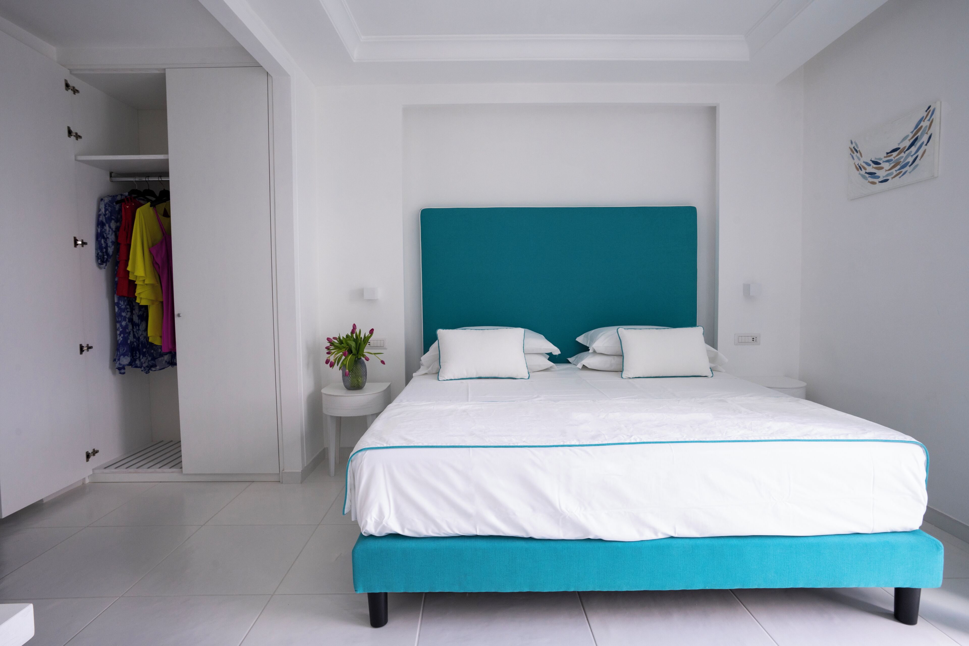 Un dormitorio con una cama, una mesita de noche, una planta en maceta y un armario con ropa de colores.