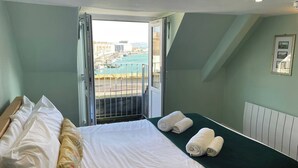 A hotel room with a bed, pillows, a view of a cityscape through a balcony, and a framed picture on the wall.
