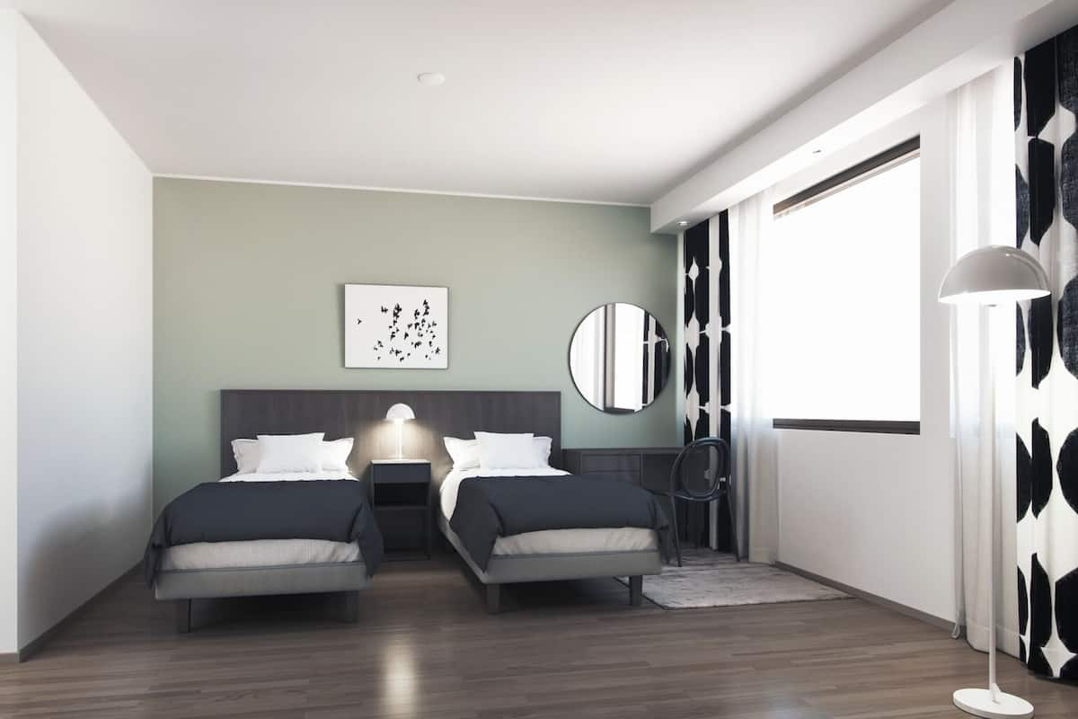 Una camera d'albergo moderna con due letti, uno specchio rotondo, un quadro a parete e un'ampia finestra.