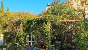 Un jardin où l’on trouve une grande variété de plantes et de fleurs, un chemin de gravier et un bâtiment surmonté d’un toit en tuiles.