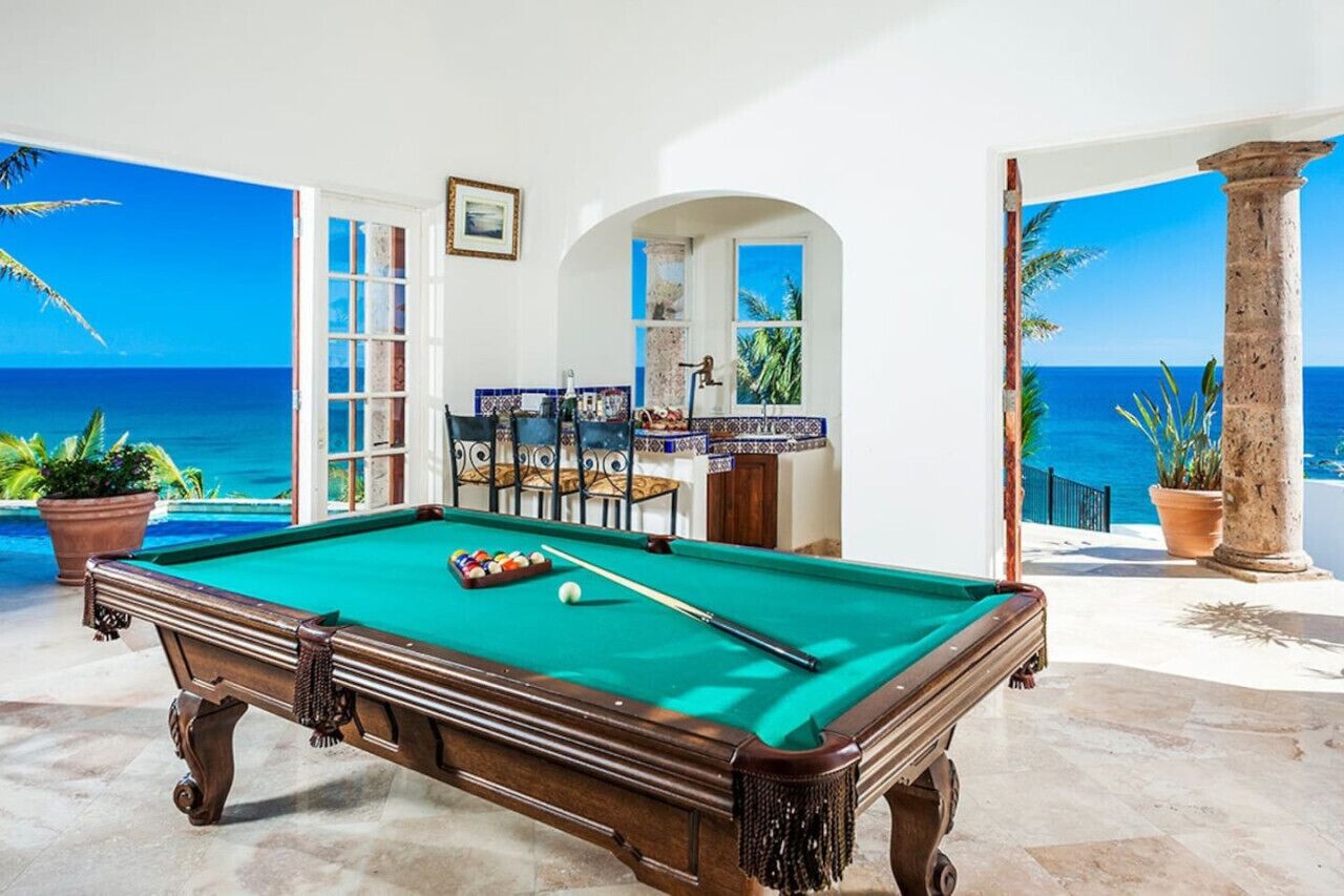 A pool table with a green felt surface, surrounded by a white interior with a view of the ocean.
