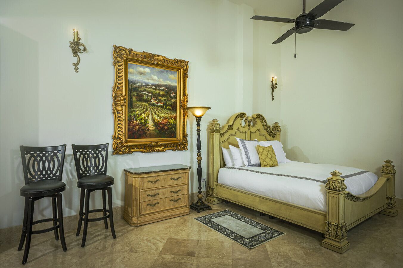 Een slaapkamer met een bed, twee stoelen, een nachtkastje, een lamp en een plafondventilator.