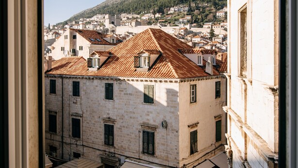 A view from a window of a building with a courtyard, featuring stone buildings with tiled roofs and a hillside in the background.