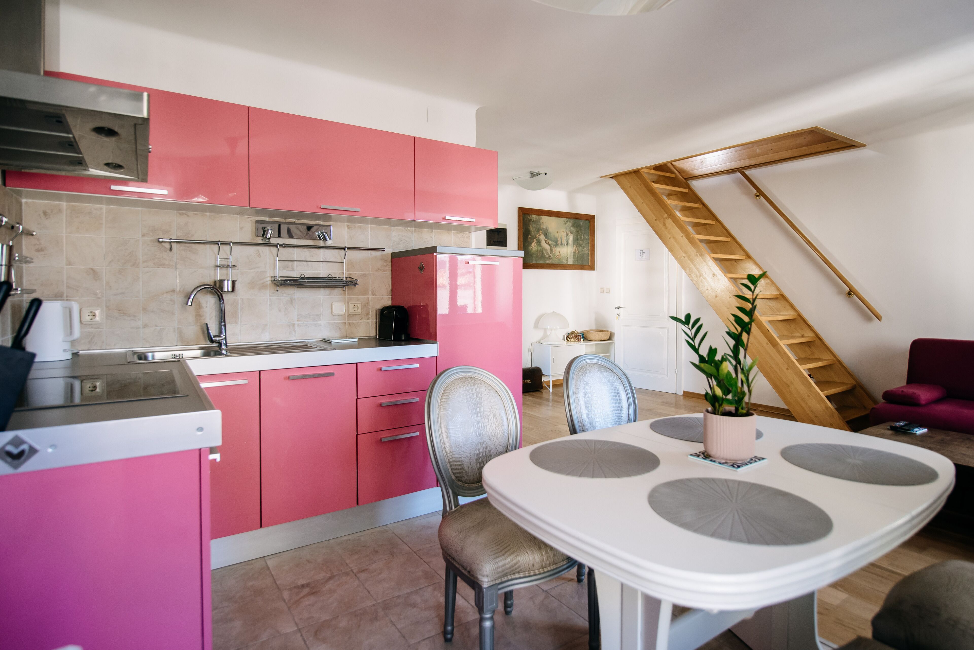 A modern kitchen with pink cabinets, a white dining table with grey accents, and a wooden staircase.
