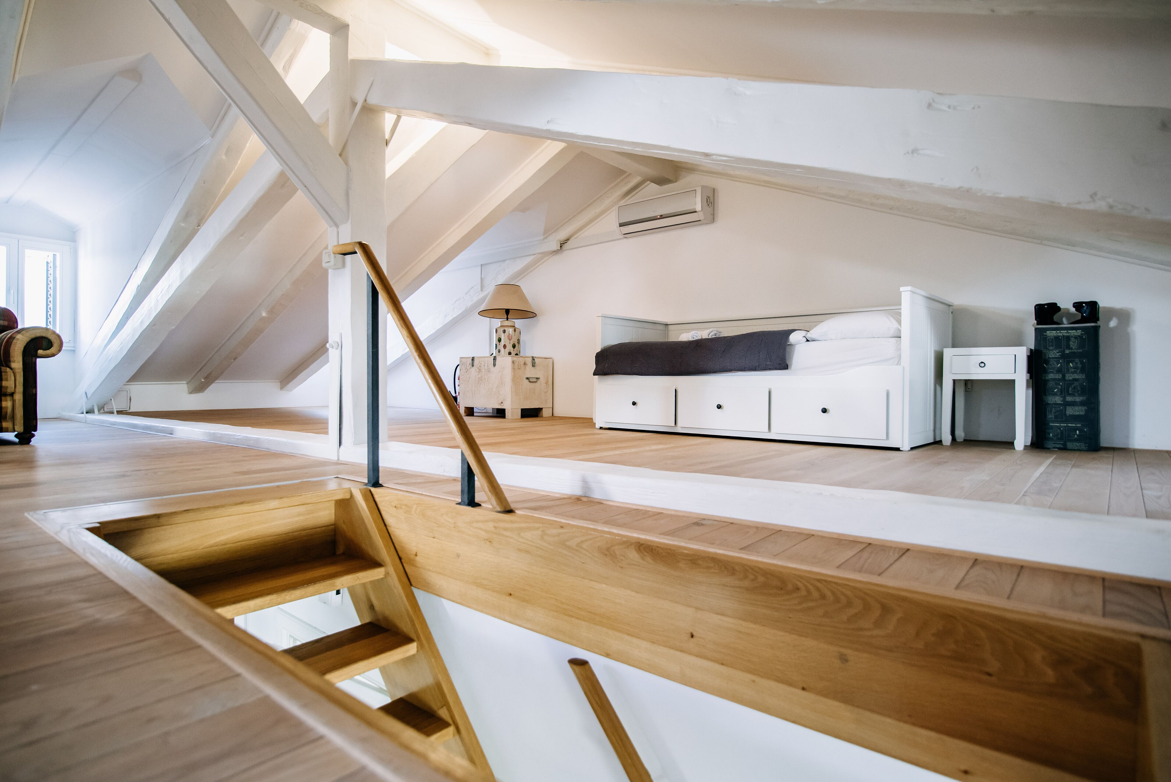 A modern loft bedroom with a wooden staircase, a bed, bedside tables, and a view of the outdoors.