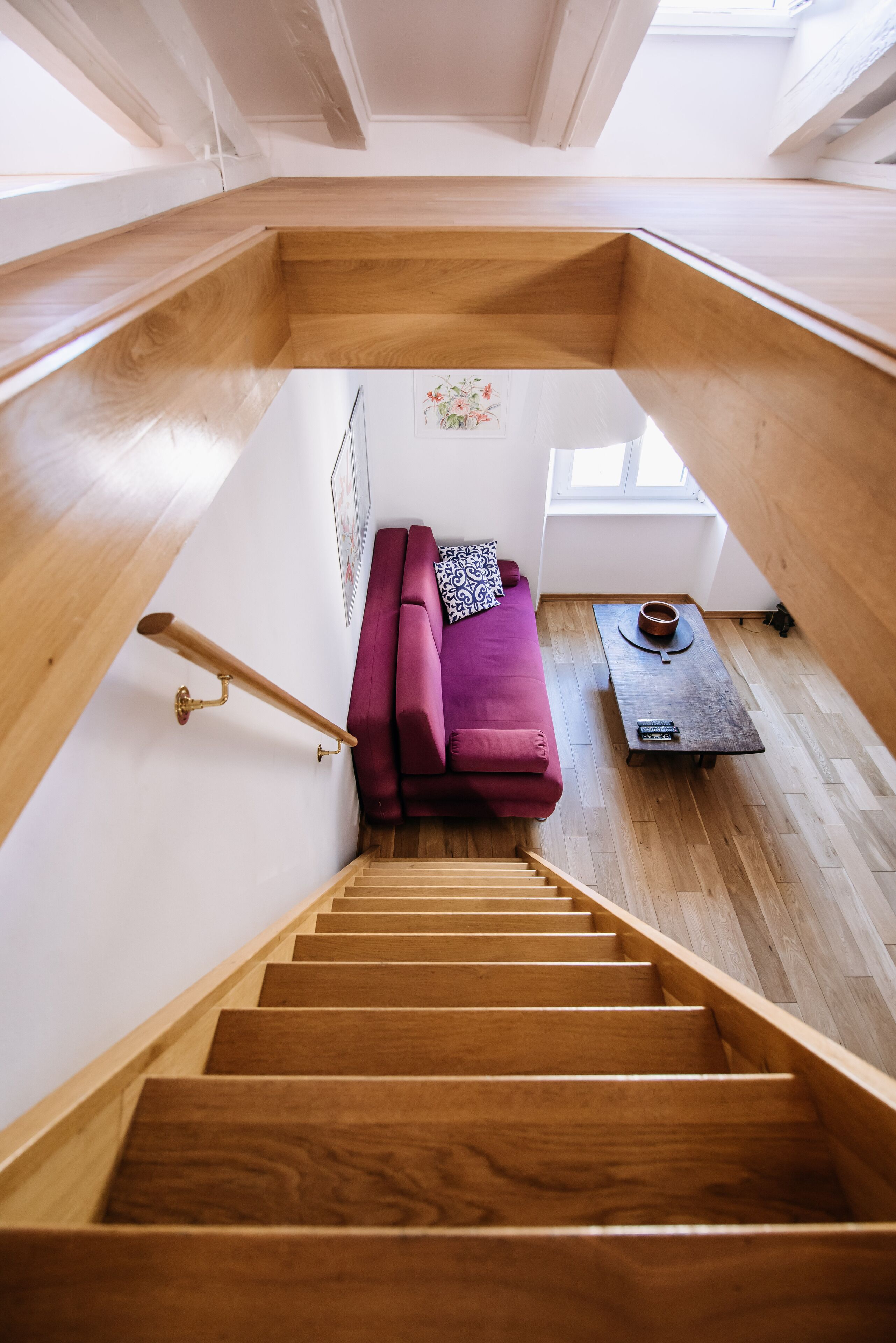 Una escalera de madera conduce a un dormitorio tipo altillo con un sofá morado y una mesita.