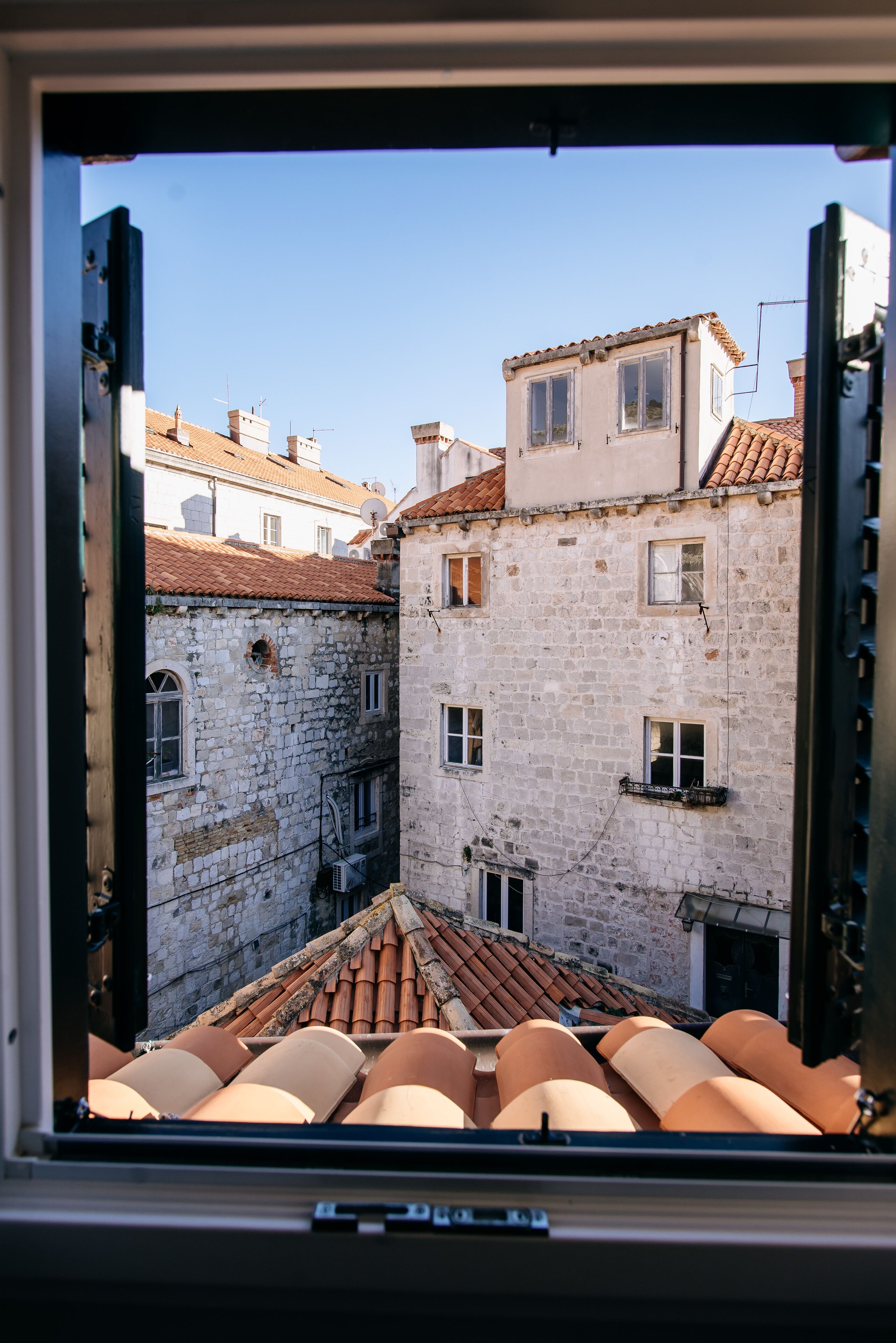 A view through a window of a narrow street with stone buildings and terracotta roof tiles.