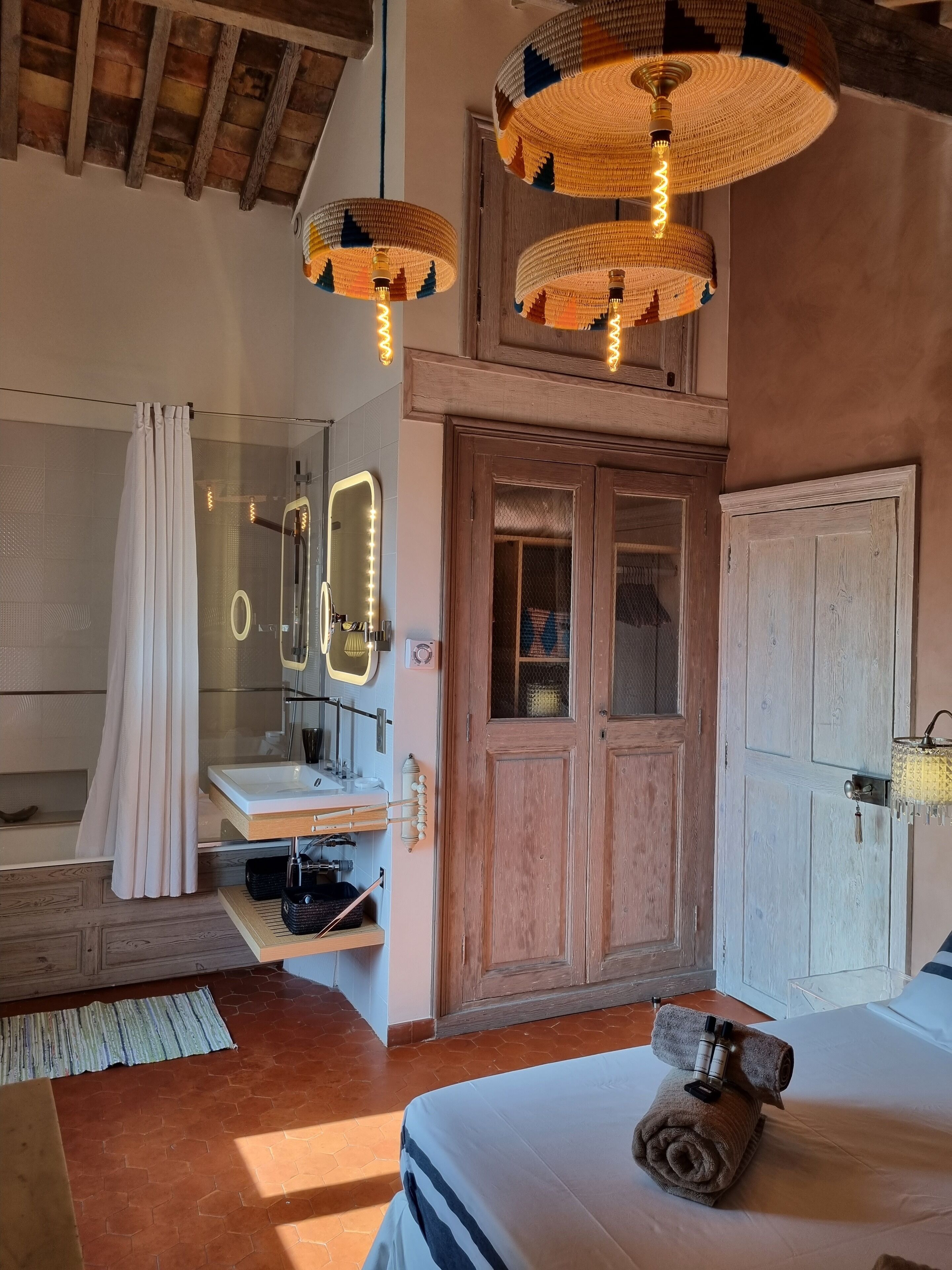 Un dormitorio con puerta de madera, una cama con toalla enrollada, un tocador con espejo e iluminación, y una lámpara colgante tejida.