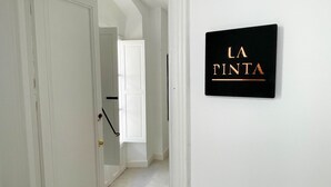 Een hal met een deur die leidt naar een kamer, een zwart bord met ‘LA TINTA’ in gouden letters en een witte deurpost.
