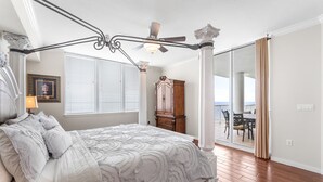 Une chambre à coucher avec un lit à baldaquin, un ventilateur de plafond, une grande fenêtre avec des stores, une armoire en bois et une vue sur l’extérieur.