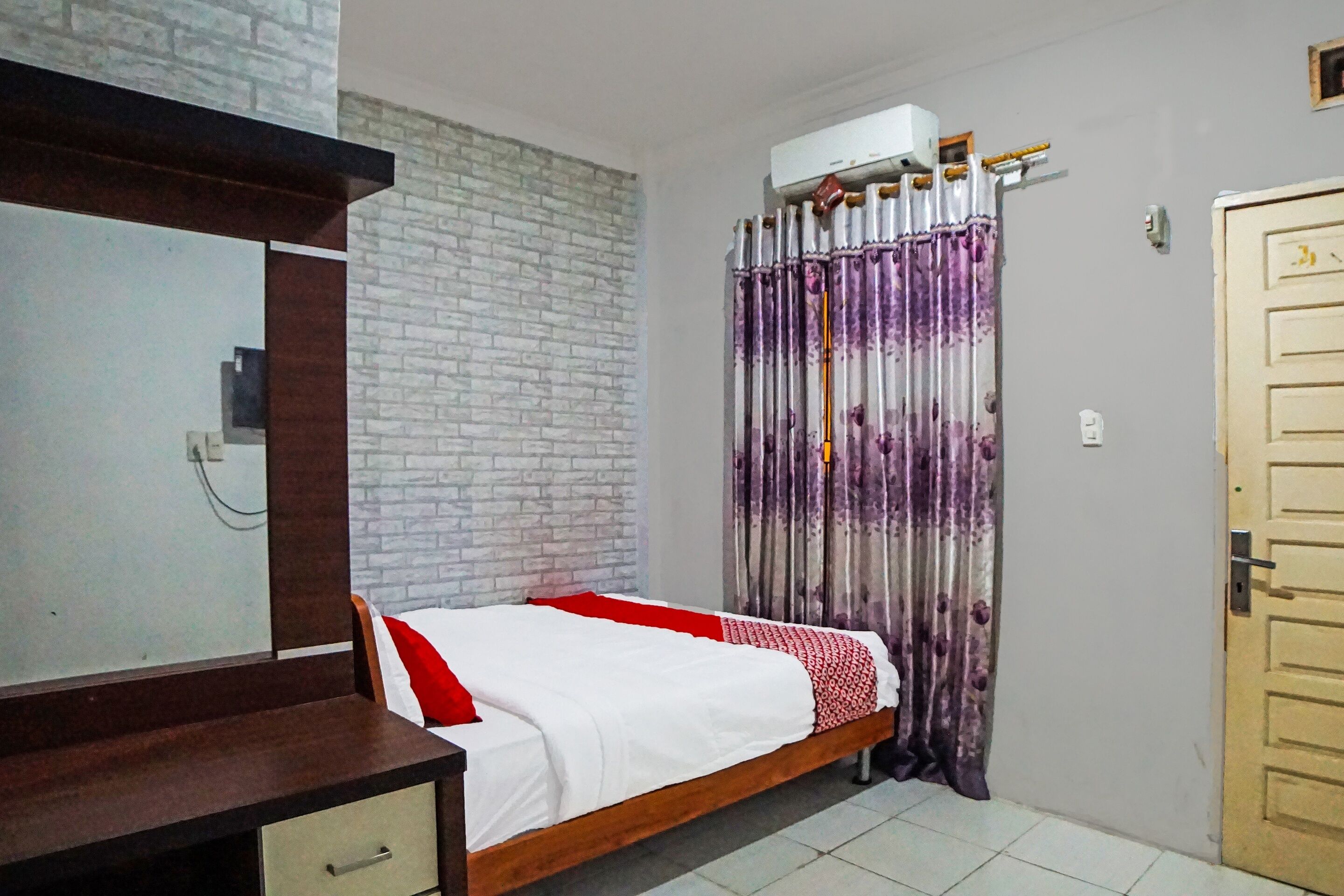 A bedroom with a bed, a wooden dresser, a brick-patterned wall, a purple and white curtain, and a door.
