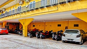 Un estacionamiento con varias motocicletas y algunos automóviles, un edificio amarillo con balcón y suelo de baldosas.