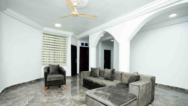 A spacious living area with a grey sectional sofa, a dark armchair, and a ceiling fan. - The Boaz Executive Place (Kumasi)