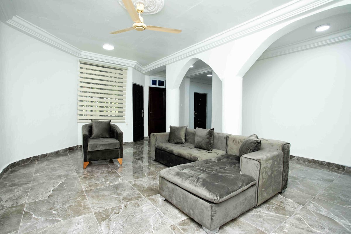 Uma sala de estar ampla com um sofá modular cinza, uma poltrona escura e um ventilador de teto.