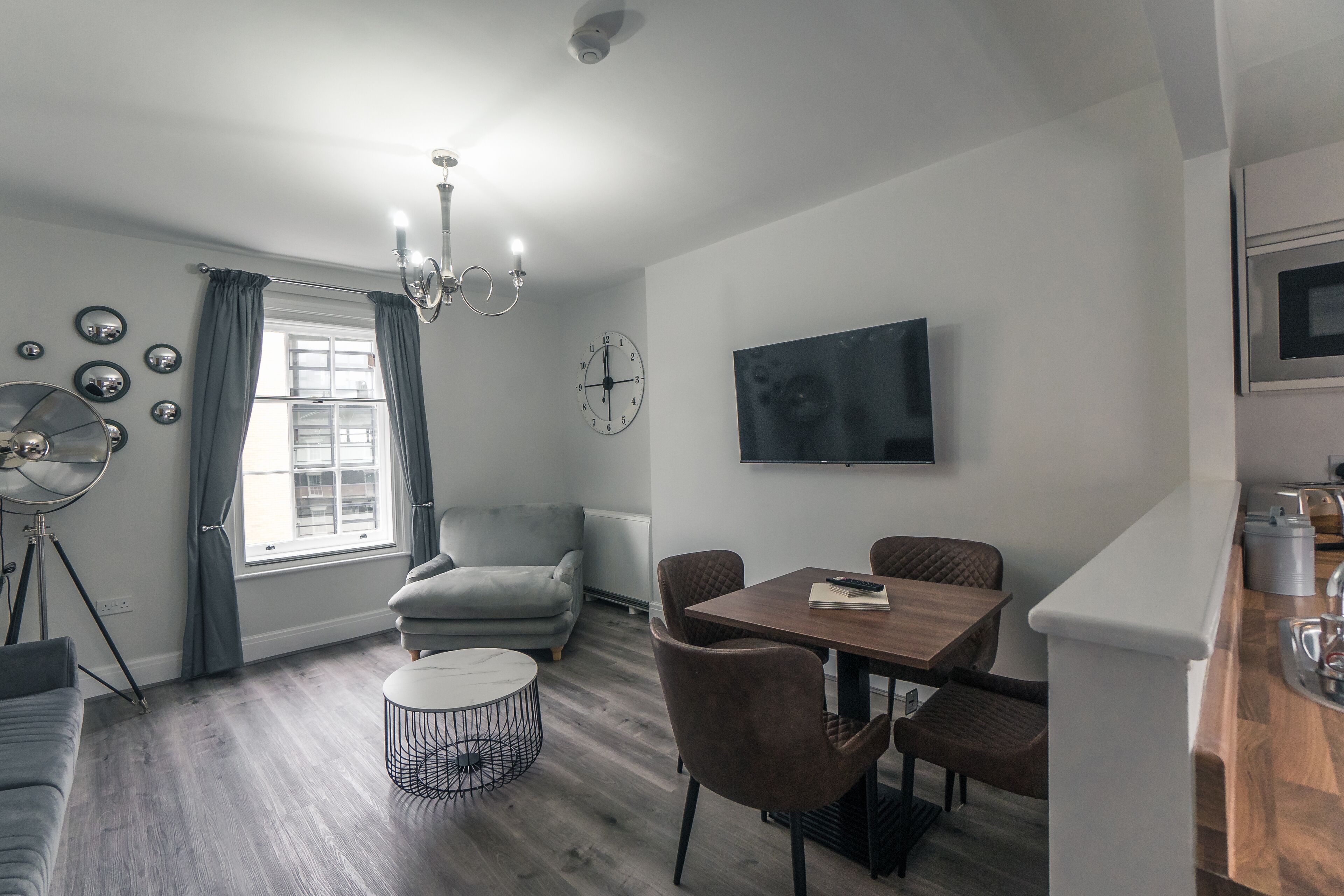 A modern living room with a dining area, grey curtains, a chandelier, a flat-screen TV, and a clock on the wall.