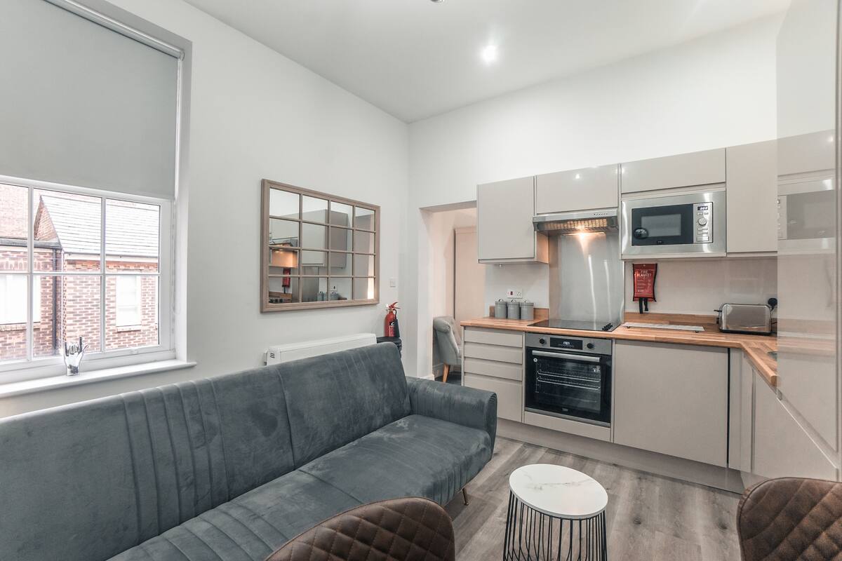 A modern kitchen with a grey sofa, a small round table, and a view of the outside through a window.