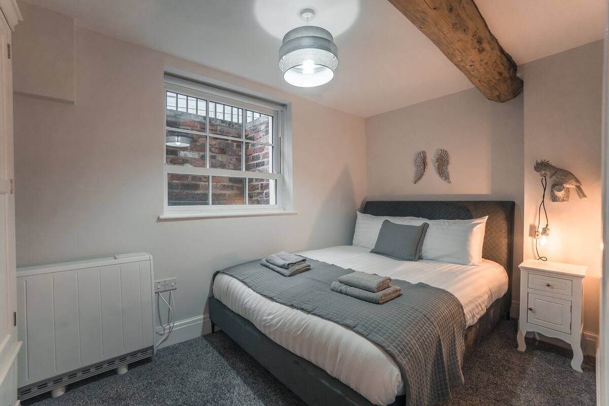 A bedroom with a bed, a nightstand, a window with a view of a brick wall, and a ceiling light.
