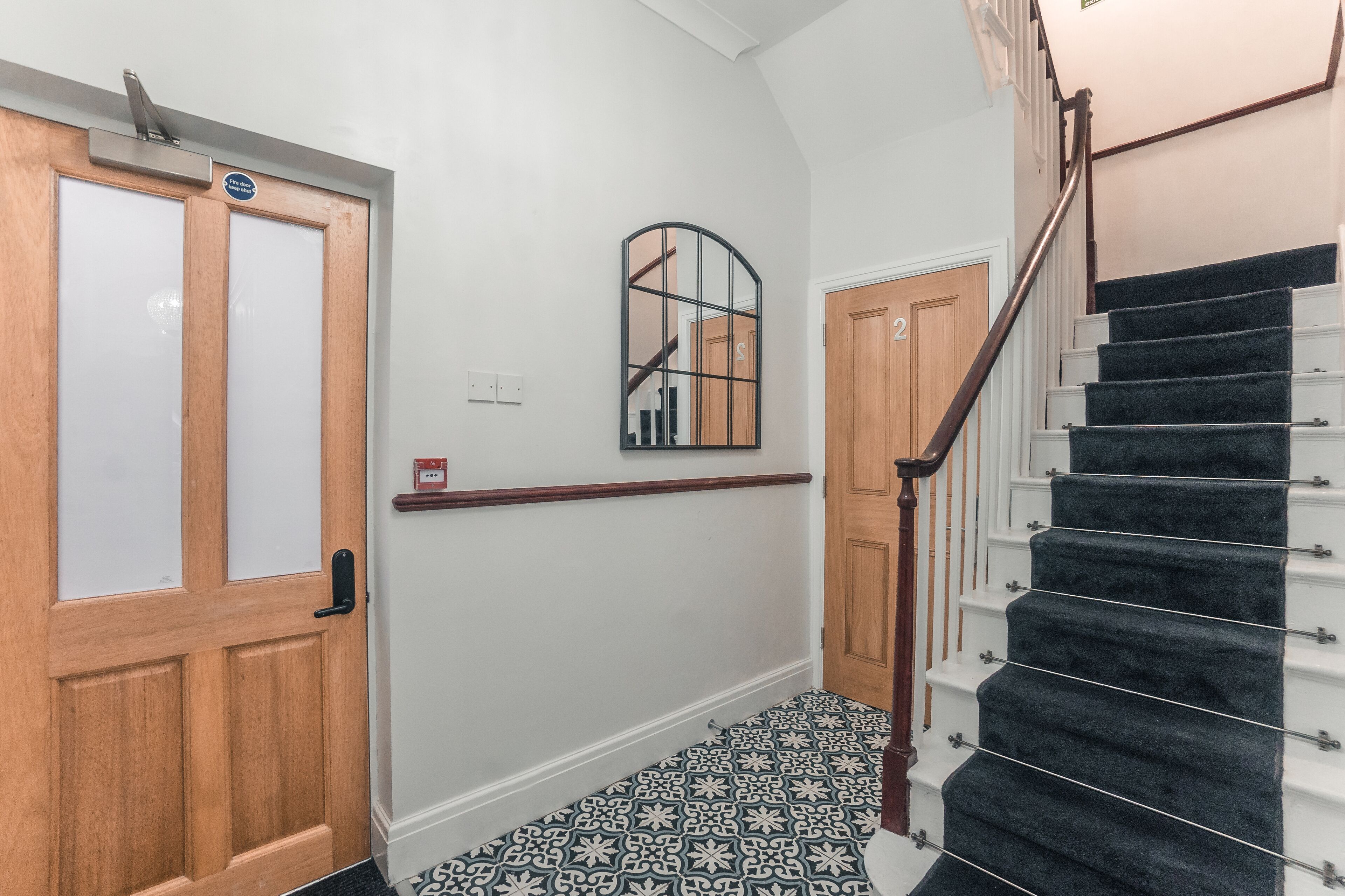 A hallway with a wooden door, a black and white patterned floor, a mirror, and a staircase with a wooden handrail.