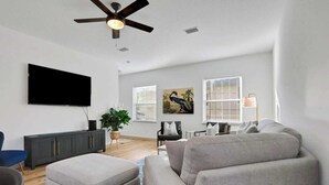 A modern living room with a flat-screen TV, grey sofas, a ceiling fan, and a large window with blinds. - New Construction in Historic Linconville (Saint Augustine)