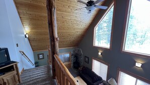 Ein gemütliches Blockhaus-Interieur mit einer Holztreppe, einem Steinkamin, einer Couch und großen Fenstern mit Blick auf den Wald.