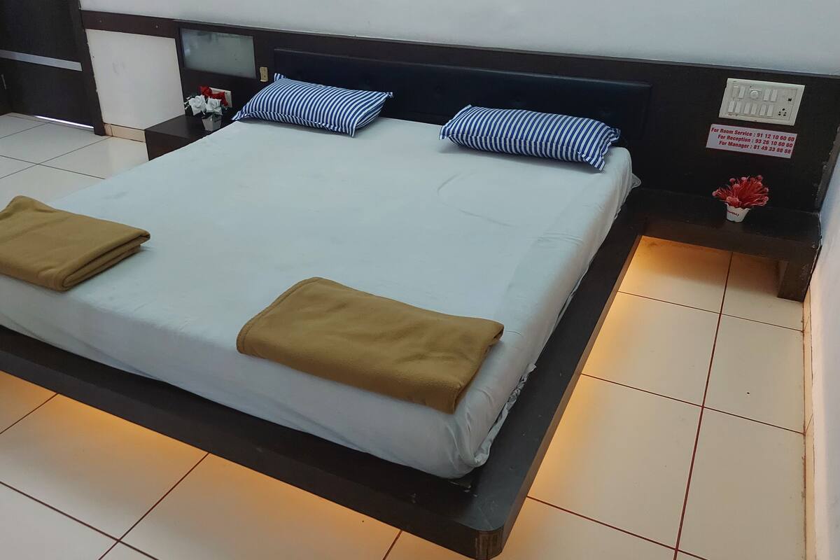 A neatly made bed with two brown blankets, a headboard, and a nightstand with a lamp and a red flower vase.