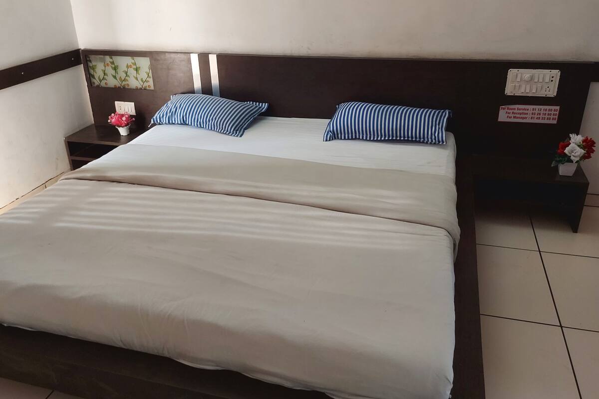 A double bed with white bedding, two striped pillows, a nightstand with a phone and a small vase of flowers, and a wall-mounted sign with room service times.