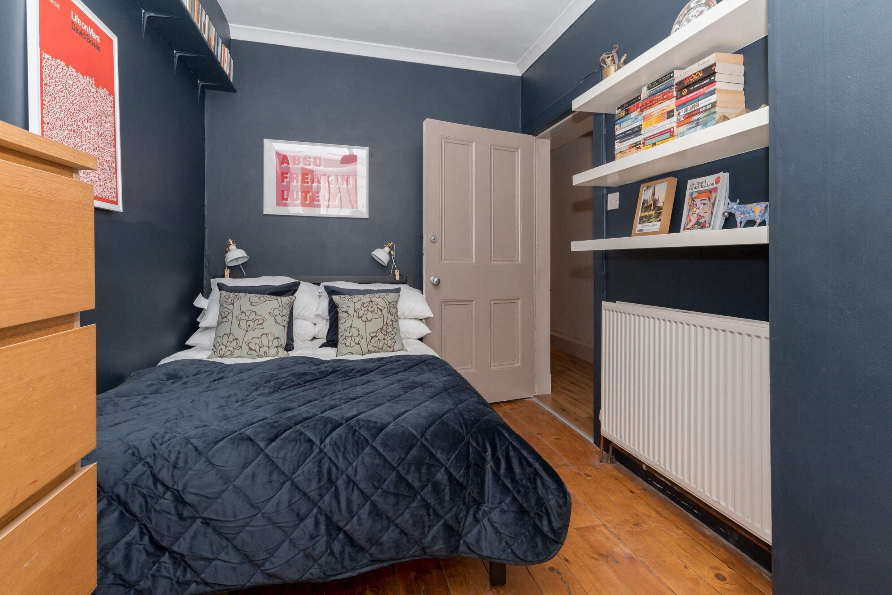 A bedroom with a dark blue wall, a bed with a quilted cover, a white radiator, and a bookshelf with books and decorative items.