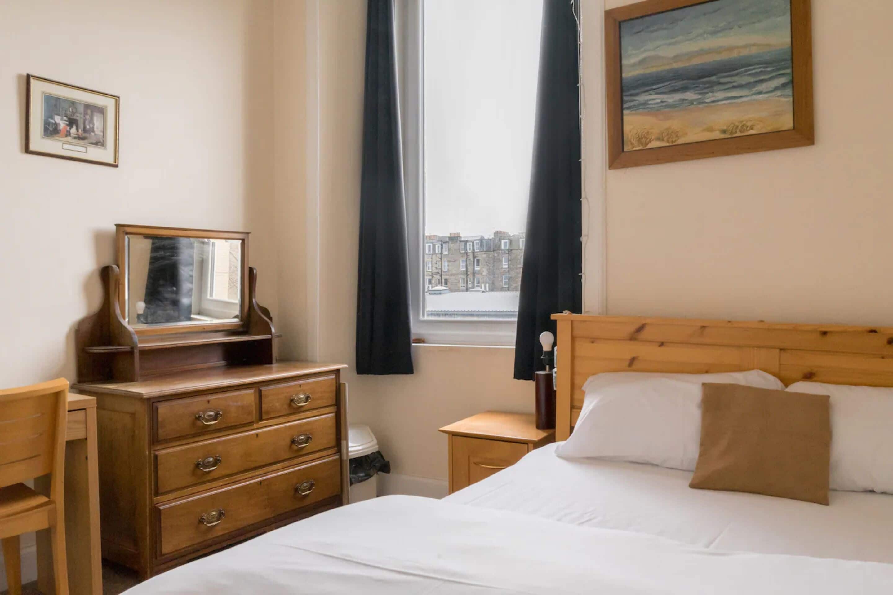 Quarto com cama de madeira, cômoda, mesa de cabeceira, janela com cortinas e um quadro na parede.