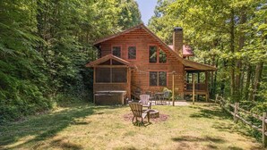 A rustic log cabin with a deck, outdoor seating, and a fire pit in a forested area.
