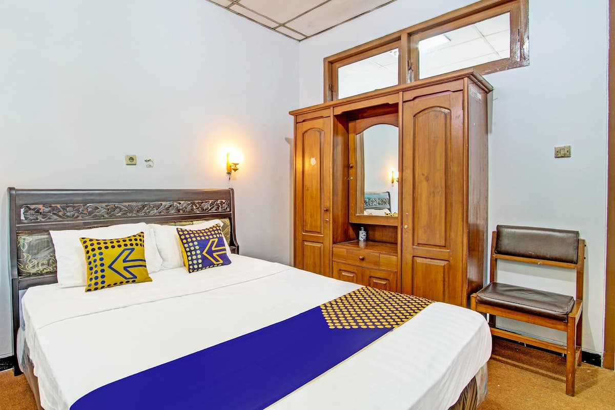 Een net opgemaakt bed met een blauwe spreideken en decoratieve kussens, een houten garderobe met een spiegel, en een enkele stoel.