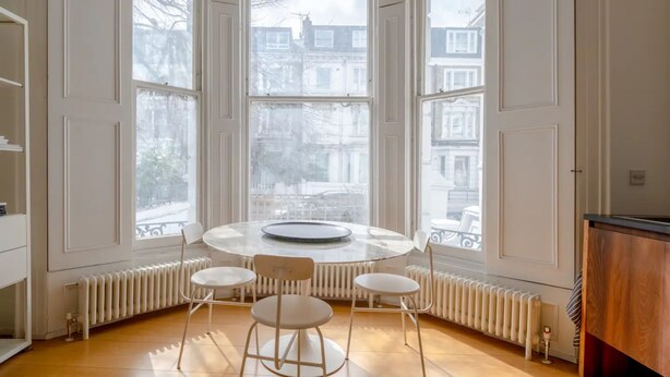 A room with a round table and four chairs, large windows with a view of buildings, and a radiator under the windows. - Stylish & Light Victorian 2BD Flat - Holland Park (London)