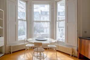 Une pièce avec une table ronde et quatre chaises, de grandes fenêtres donnant sur des immeubles, et un radiateur placé sous les fenêtres.