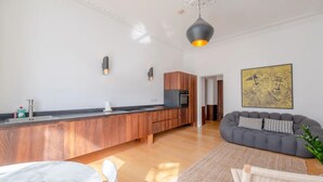 A modern kitchen with wooden cabinets, a marble countertop, and a built-in oven. There is a gray sofa and a woven rug in the living area. - Stylish & Light Victorian 2BD Flat - Holland Park (London)