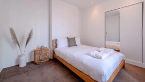 A modern bedroom with a wooden bed, white bedding, a large mirror, and a wicker basket. - Stylish & Light Victorian 2BD Flat - Holland Park (London)