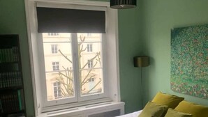 Une chambre avec une grande fenêtre, un lit avec une literie blanche et verte, une bibliothèque et une lampe fixée au mur.