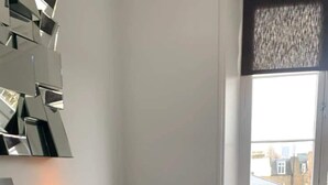 Un salon moderne avec une cheminée, une table ronde accompagnée de deux chaises noires, un vase jaune et une fenêtre donnant sur des immeubles.