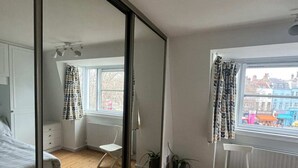 Quarto com um espelho grande, piso de madeira, uma cama, janela com cortinas e vista para edifícios.