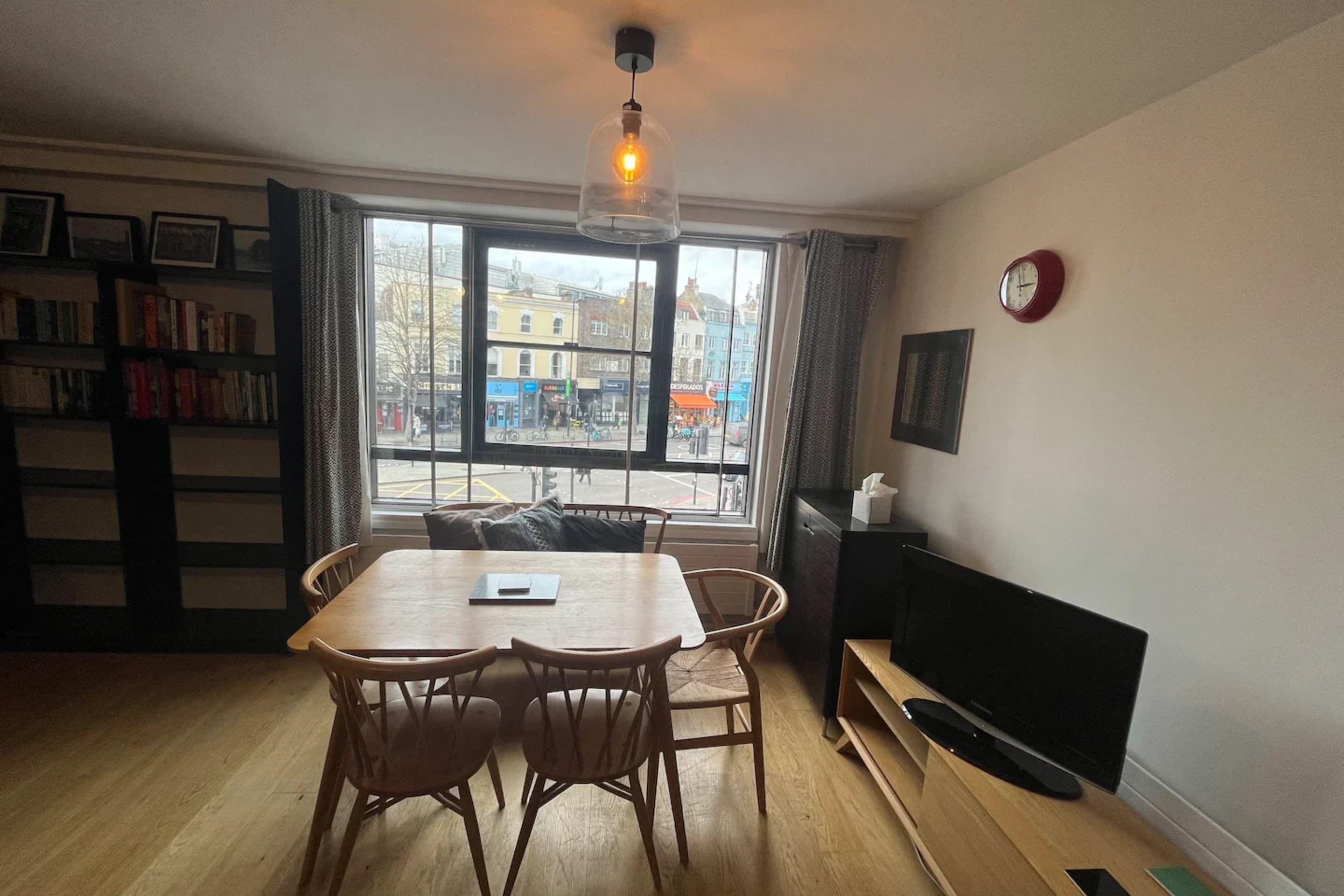 Een gezellige woonkamer met een houten eettafel, twee stoelen, een televisie, een boekenkast en een groot raam met uitzicht over de stad.