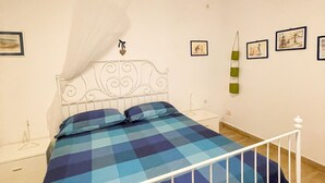 A bedroom with a bed, white metal headboard, and a checkered bedspread. There are bedside tables, a hanging lamp, and framed pictures on the wall.