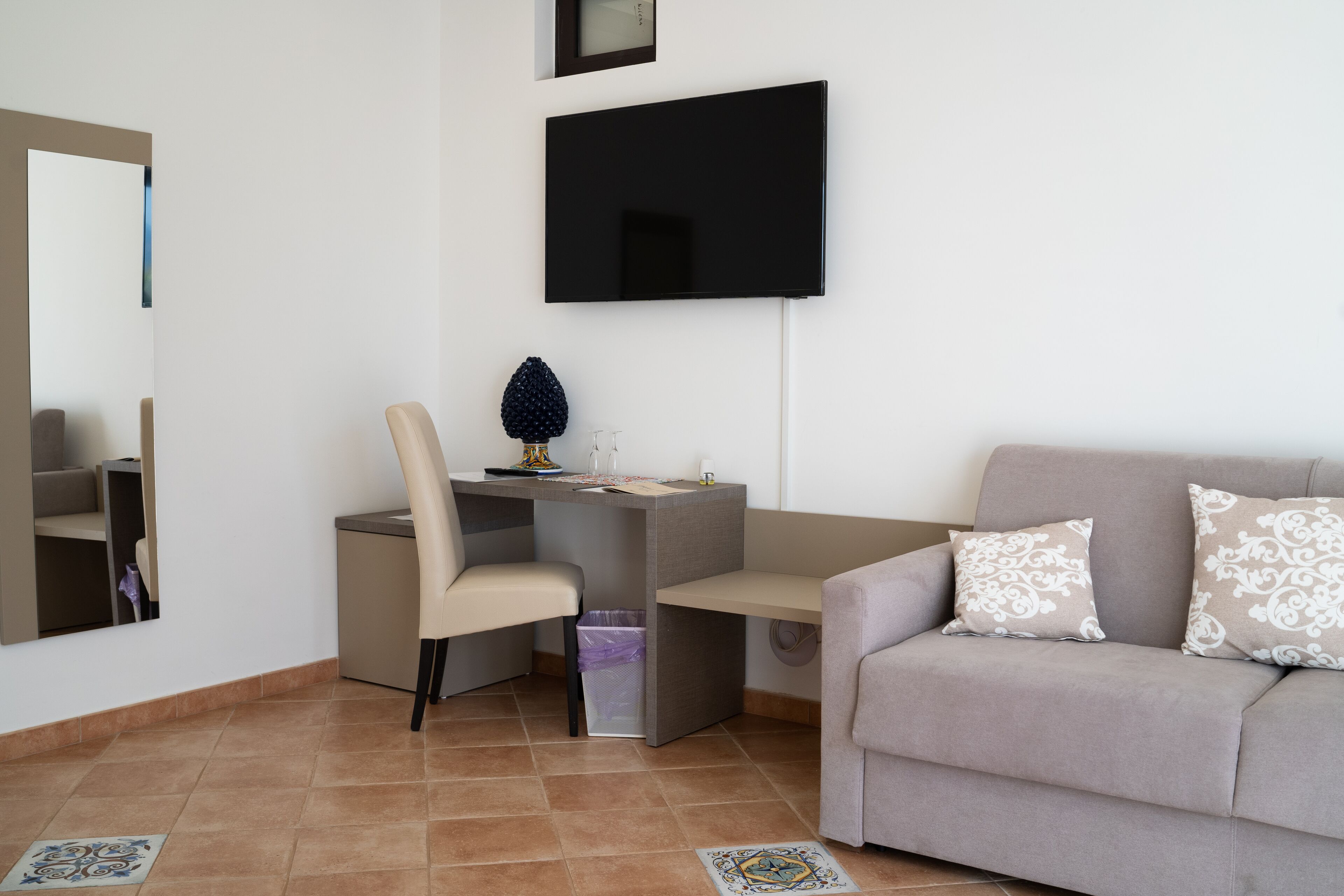 Una sala de estar moderna con un televisor de pantalla plana montado en la pared, un sofá gris con cojines estampados, un escritorio pequeño con una silla y un jarrón decorativo.