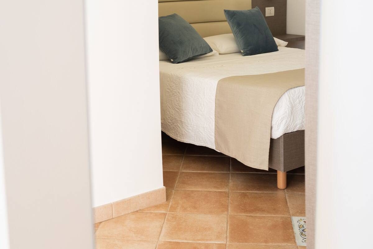 Ein Hotelzimmer mit Bett, zwei Kissen, einem Nachttisch, einem an der Wand befestigten Thermostat und einem Gemälde einer Küstenlandschaft.