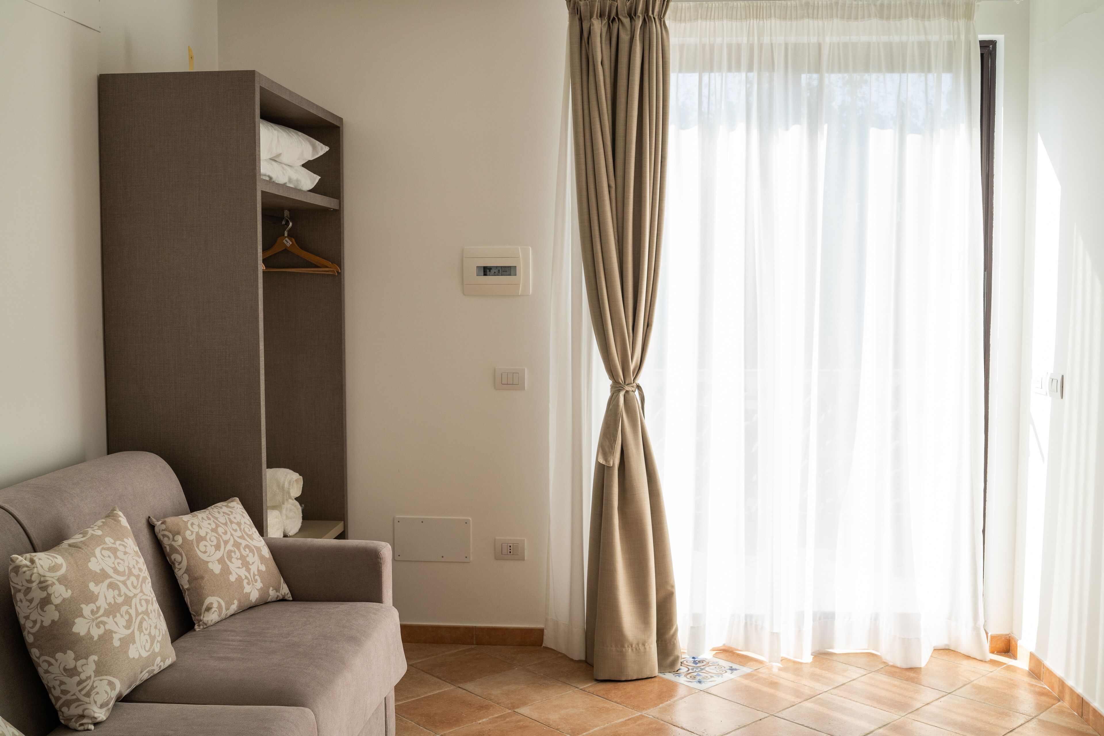 A room with a sofa, a wardrobe, a hanging light fixture, and a window with sheer curtains.