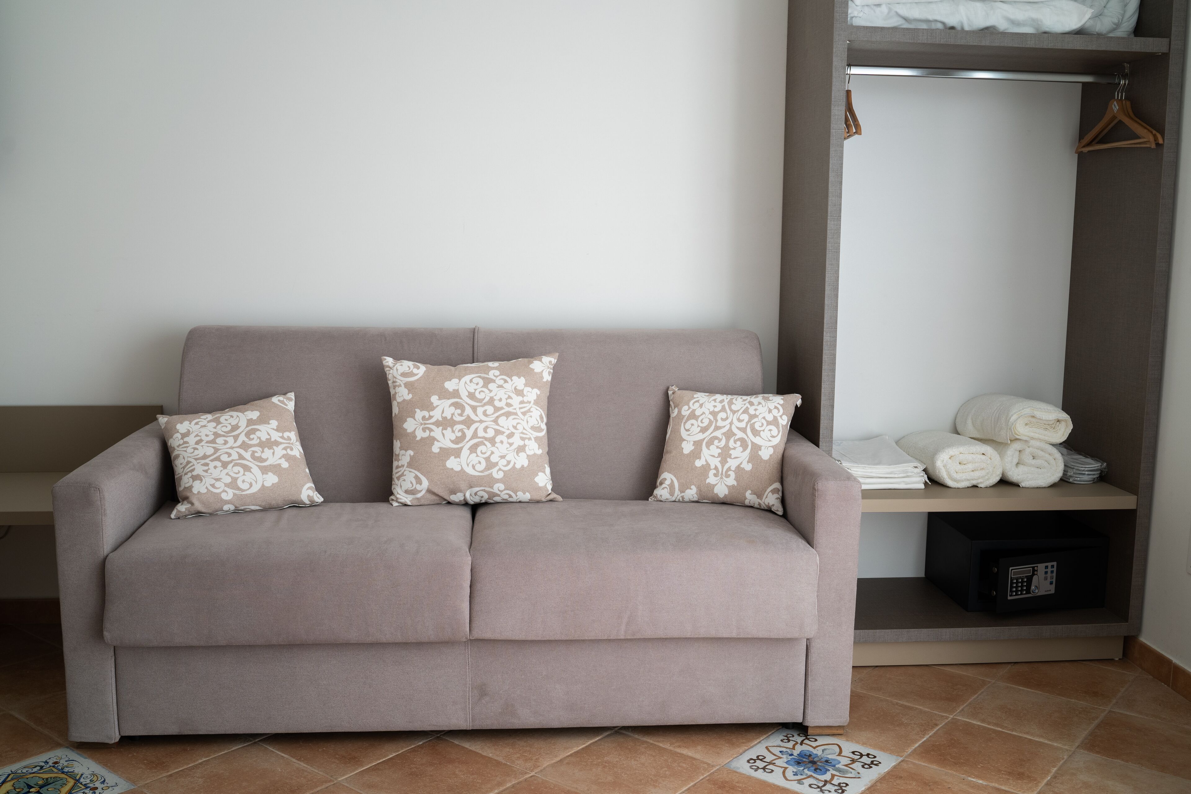 En beige sofa med mønstrede puder, en reol med foldede håndklæder og en lille, sort apparat.