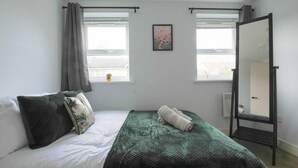 Een slaapkamer met een bed, een groene deken, een opgerolde handdoek, een spiegel en twee ramen met gordijnen.