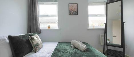 A bedroom with a bed, a green blanket, a rolled-up towel, a mirror, and two windows with curtains.