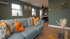 Een moderne woonkamer met een blauwe bank, meerdere kussens, een eethoek met een tafel en stoelen, en een keuken op de achtergrond.