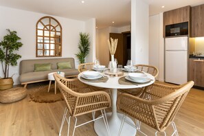 Una sala de estar moderna con una mesa de comedor redonda, sillas de mimbre, un sofá y una zona de cocina con nevera y microondas.