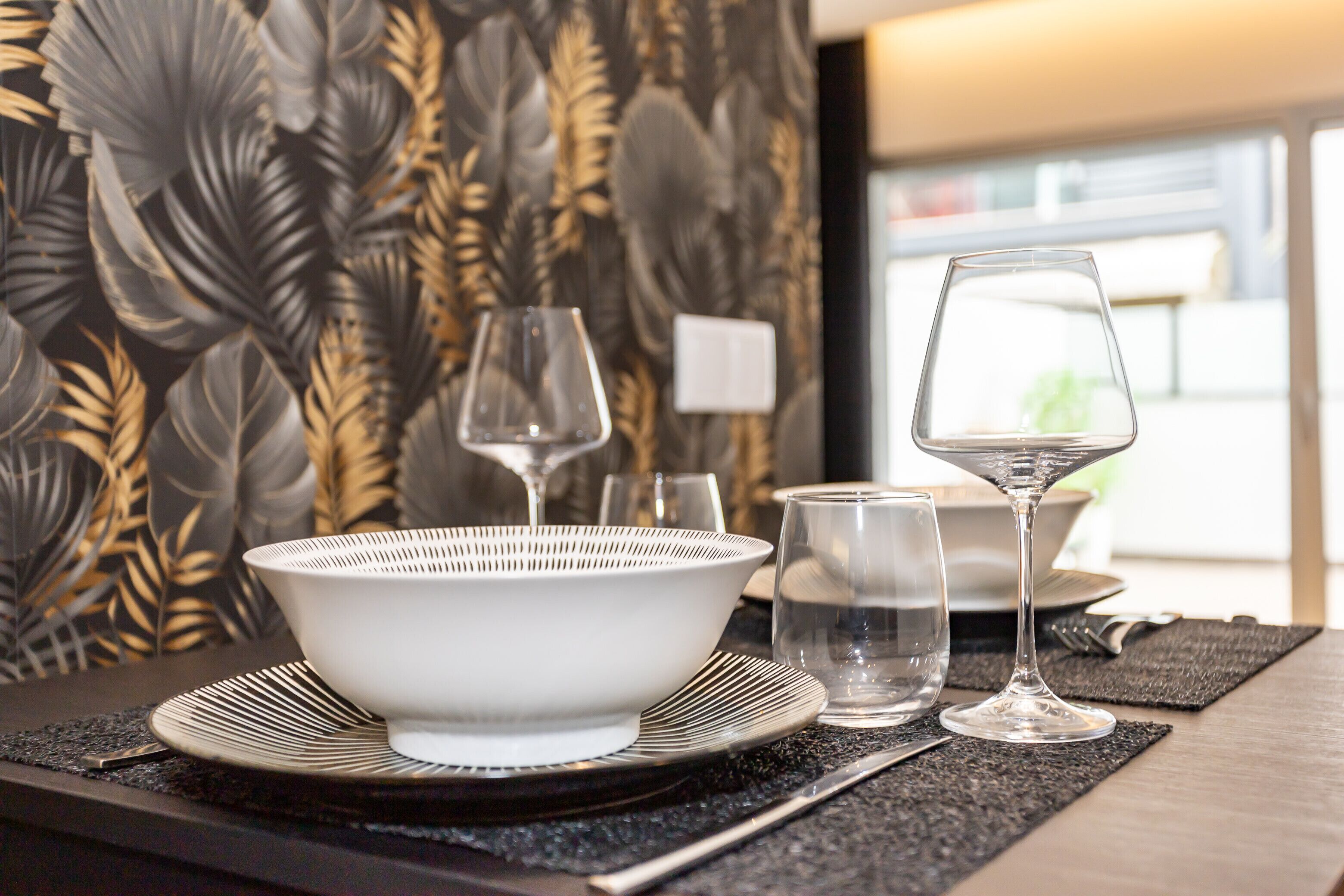 A dining table set with a white bowl, two wine glasses, and a water glass on a black placemat.